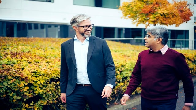 Two professionals walking outside an office building with autumn foliage in the background, discussing business topics